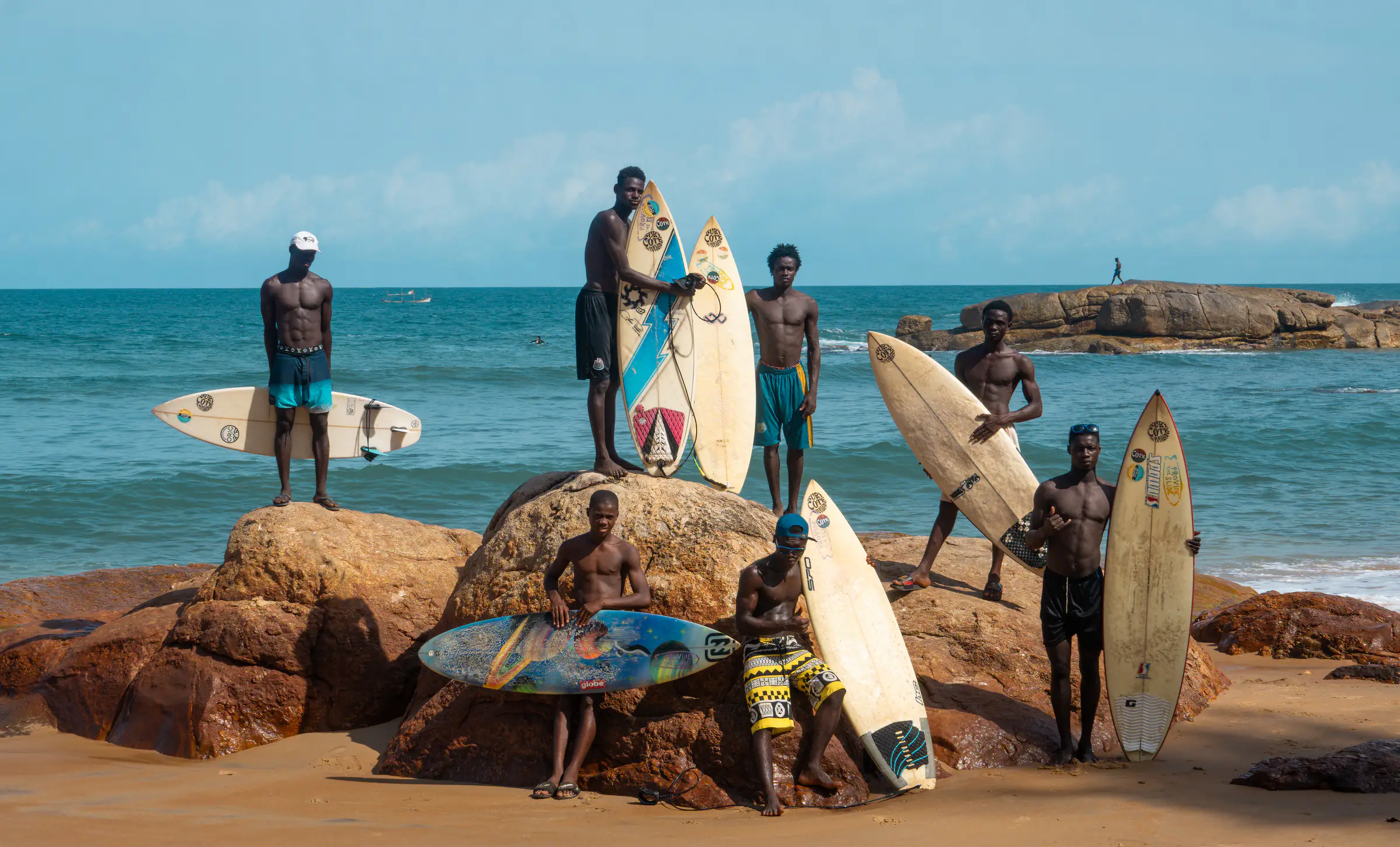 De jeunes surfeurs sur les rochers d’une plage ivoirienne, planche à la main, la mer derrière eux.