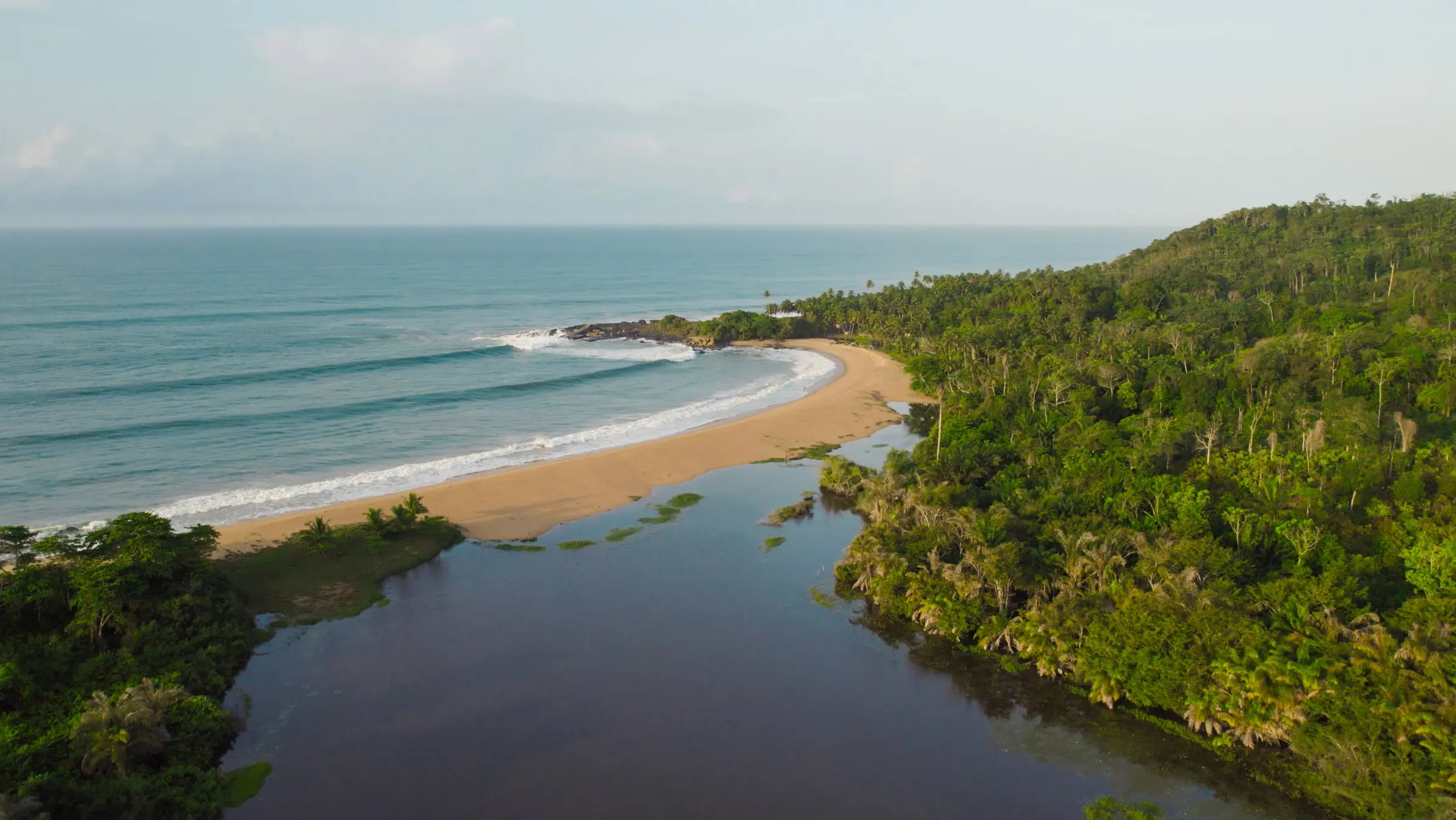 Vue aérienne d’une lagune qui rejoint la mer, une plage en croissant et une forêt dense.