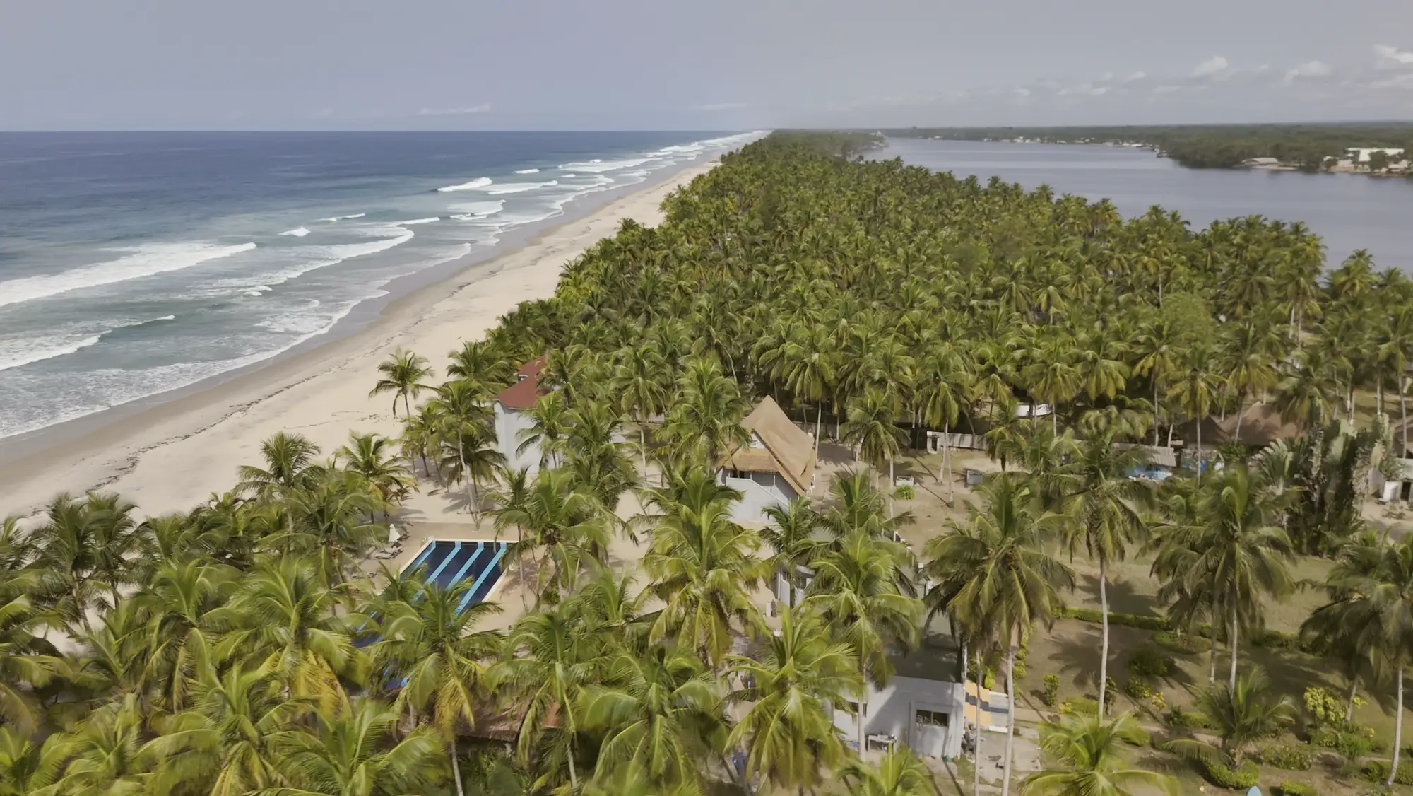 Le camp vu du ciel : cocotiers et toits de paille sur une bande de sable, l’Atlantique qui déferle à gauche, la lagune Aby derrière.