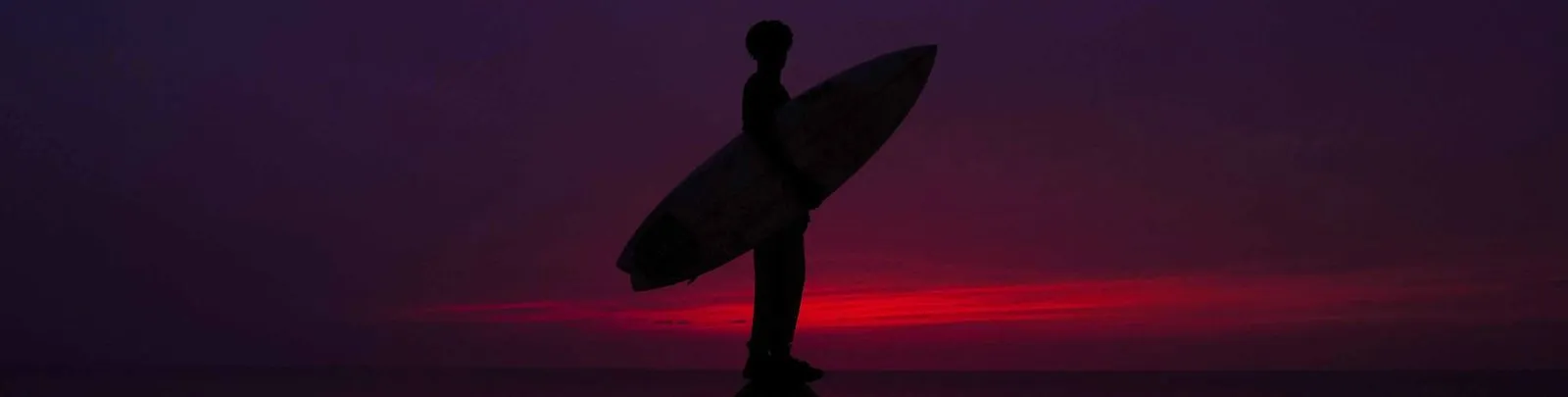 A surfer in silhouette holding his board, against a red sky at dusk.