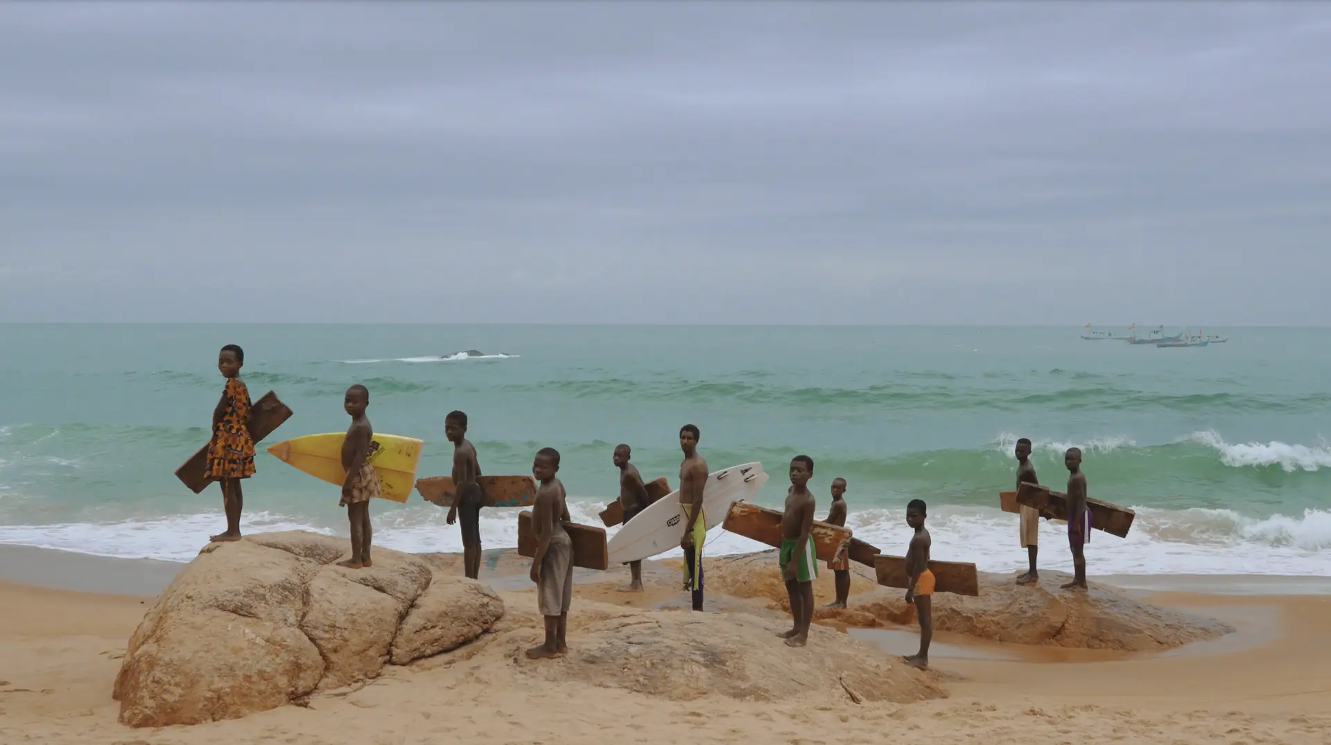 Children on the rocks of an Ivorian beach, holding wooden boards and surfboards.