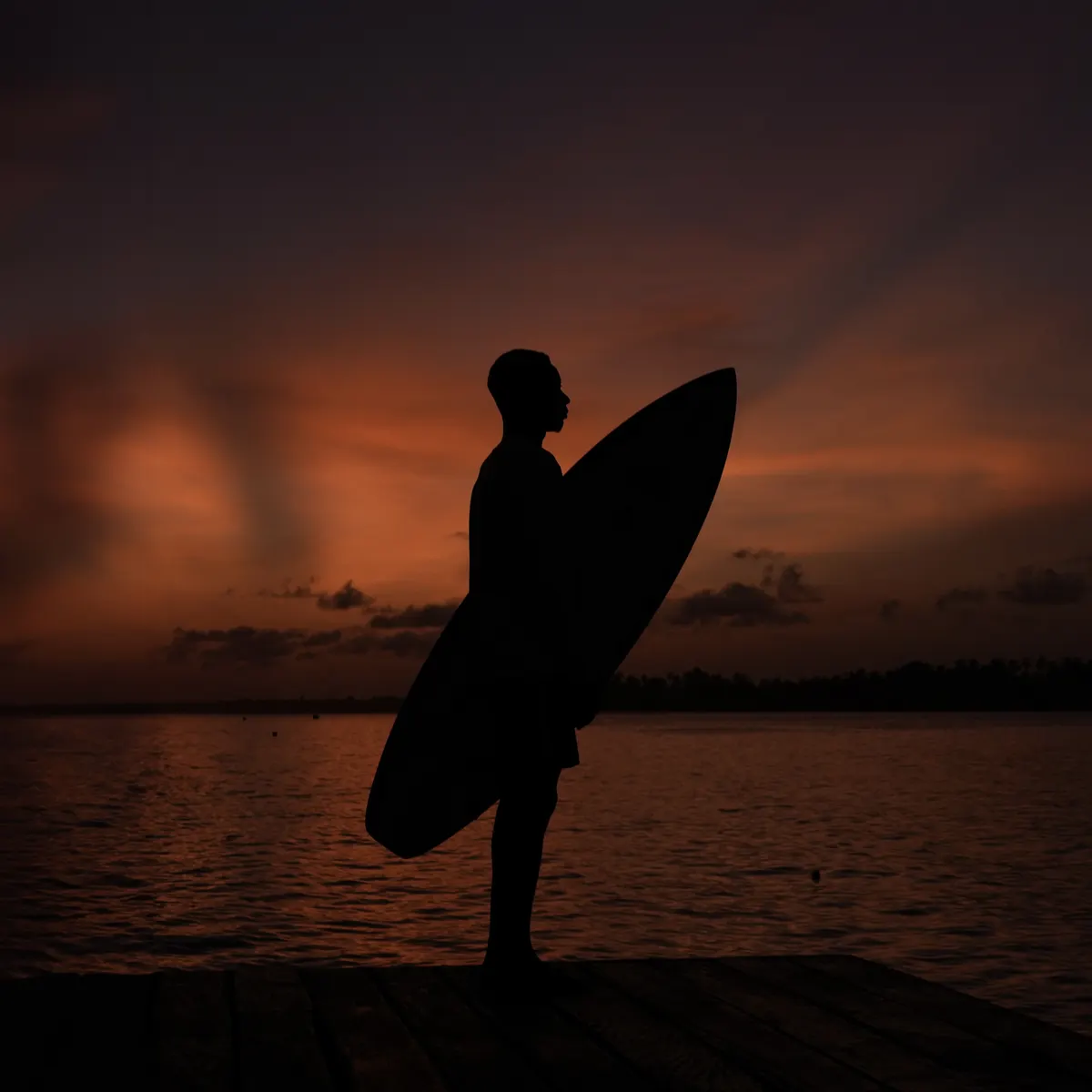 A surfer in silhouette holding his board against an orange sunset.