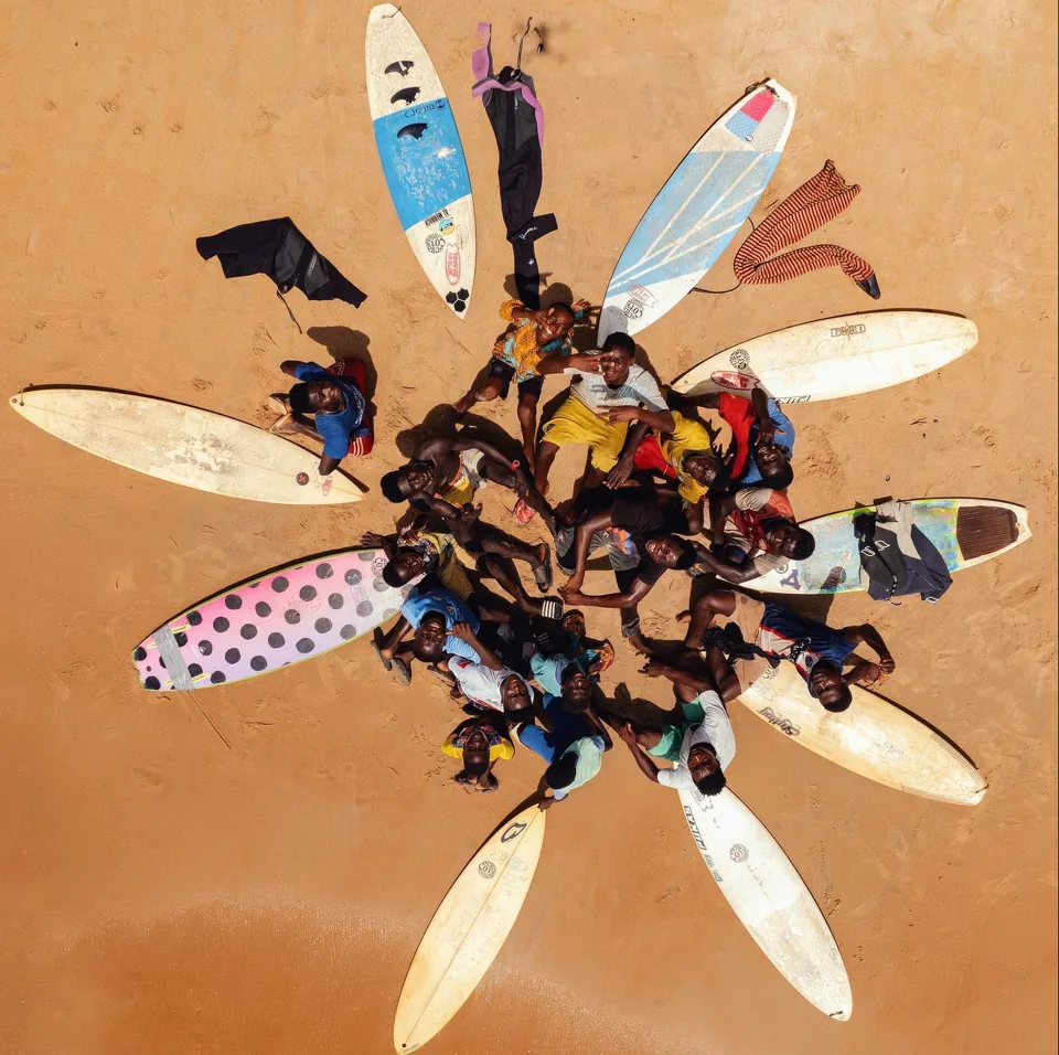 Vue du ciel : des enfants et de jeunes surfeurs allongés en cercle sur le sable, les planches autour d’eux comme des pétales.