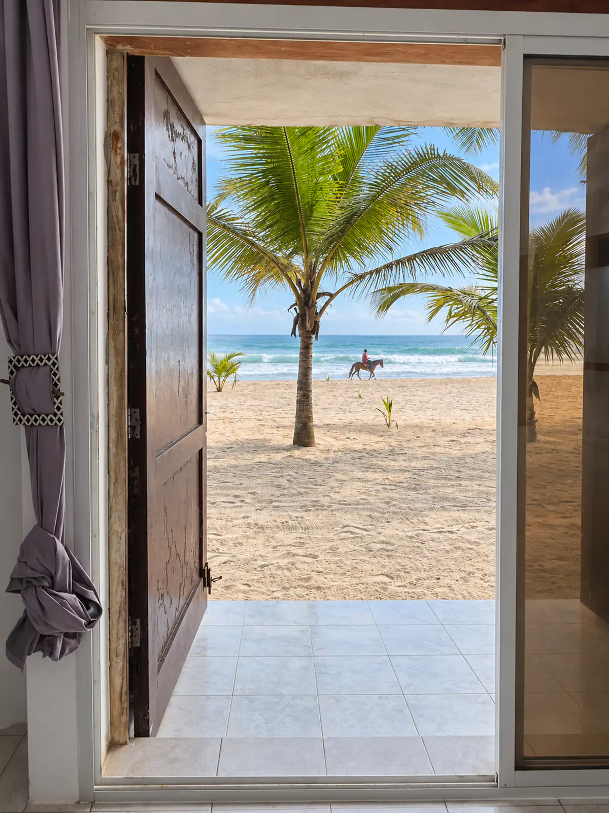 The room’s door open onto the beach: palm trees, the sea and a rider on horseback.