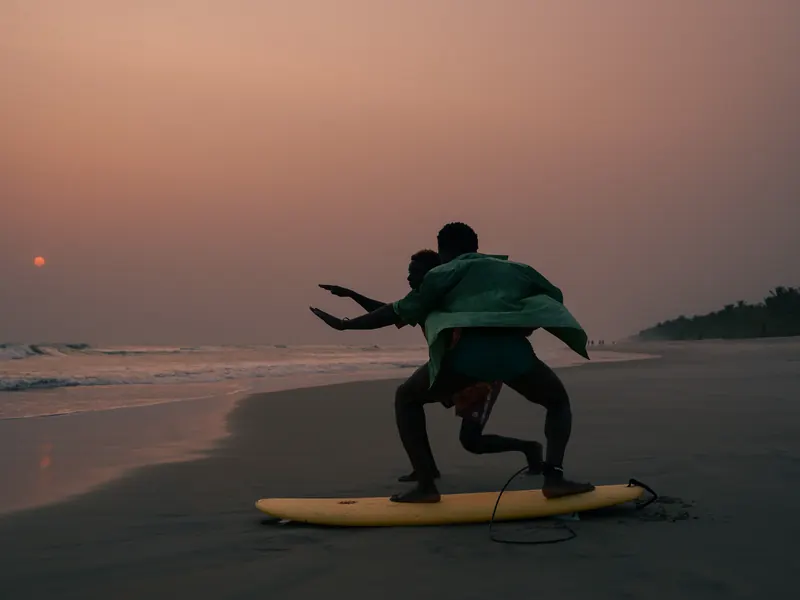 A coach showing a young surfer the stance on a board on the sand, at sunset.